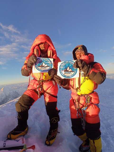 Two mountaineers in red suits holding a sign at a high-altitude summit