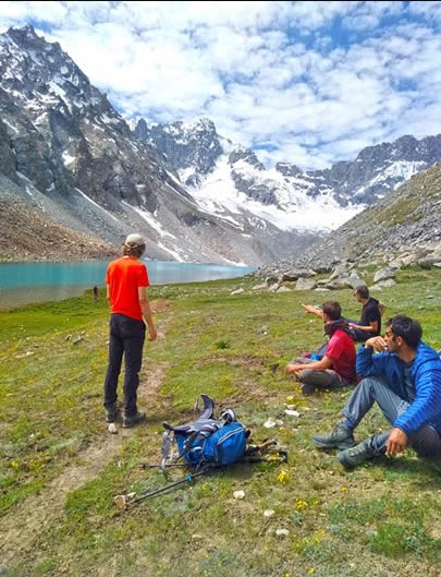 A group of trekkers resting near the beautiful green meadows and Lake, surrounded by towering peaks.